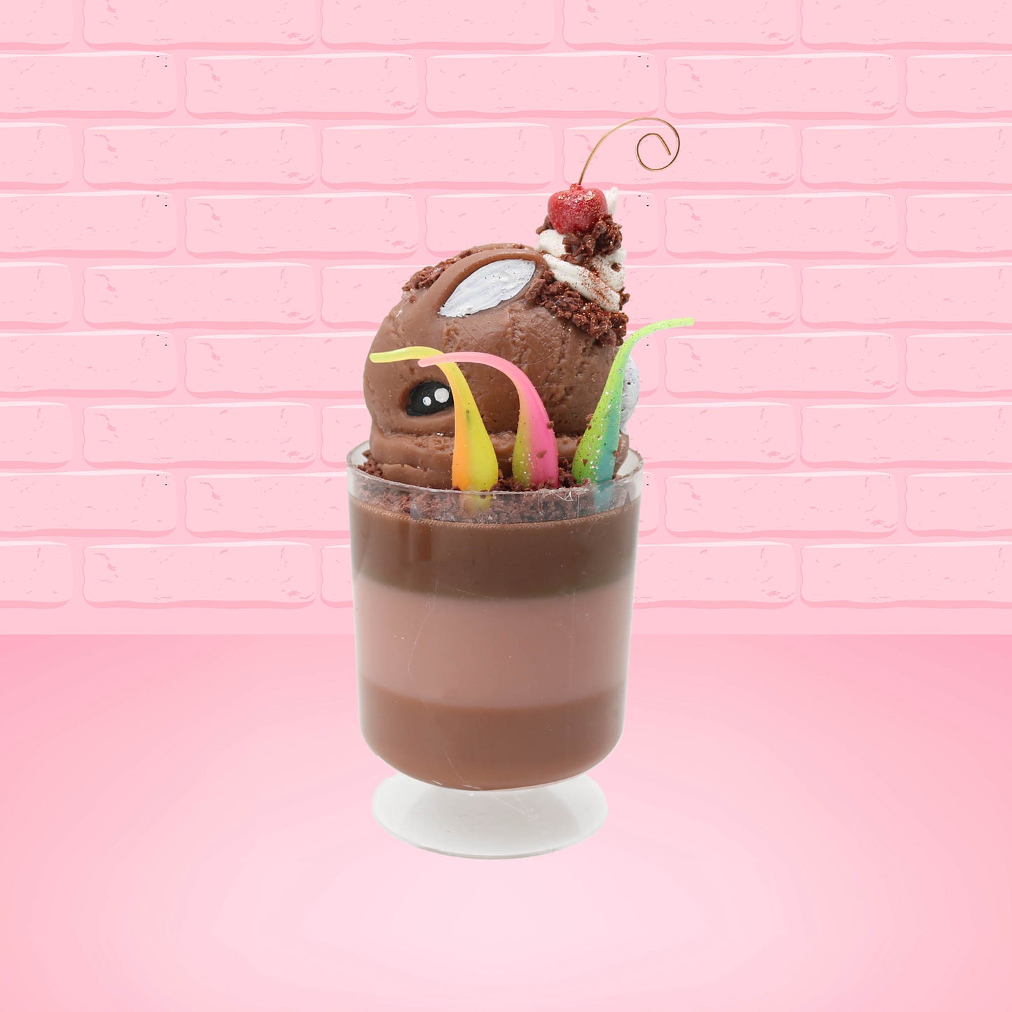 Cute kawaii bunny tall brown and light brown ice cream sundae with brown chocolate crumbles over the bunny, with a cherry on top of the whipped cream swirl.  Three bendy gummy worms are in the dish on the side. This view is from the side.