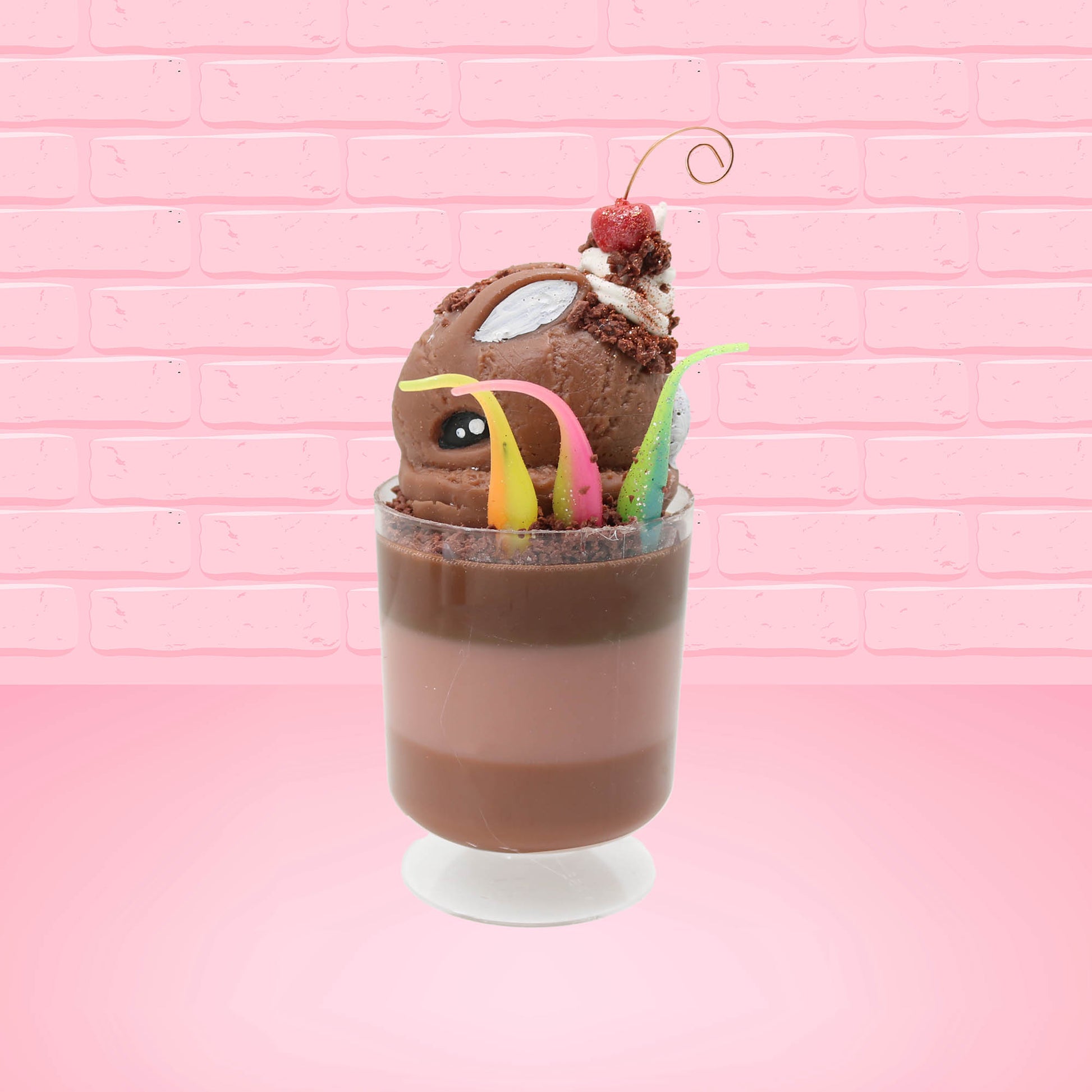Cute kawaii bunny tall brown and light brown ice cream sundae with brown chocolate crumbles over the bunny, with a cherry on top of the whipped cream swirl.  Three bendy gummy worms are in the dish on the side. This view is from the side.