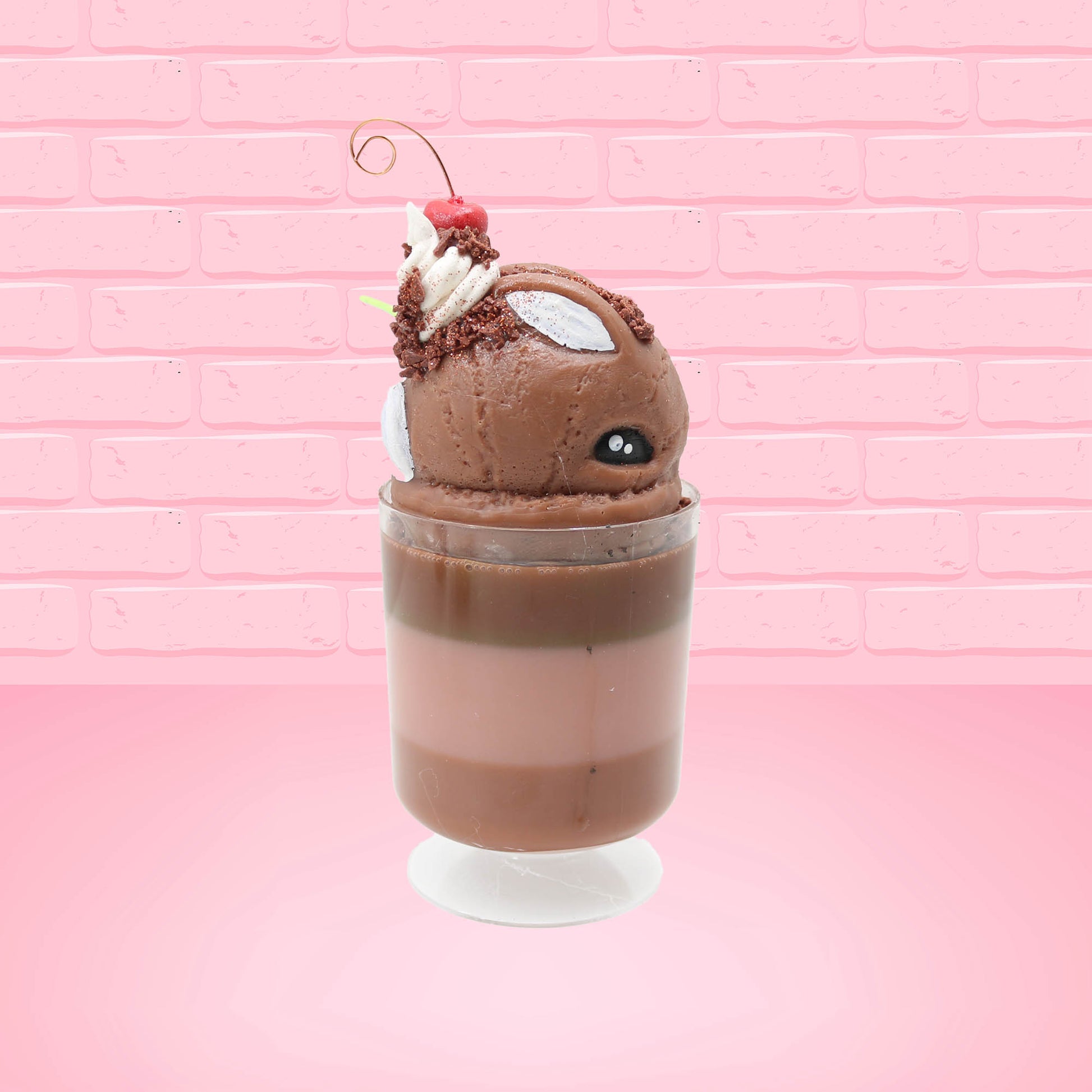 Cute kawaii bunny tall brown and light brown ice cream sundae with brown chocolate crumbles over the bunny, with a cherry on top of the whipped cream swirl.  Three bendy gummy worms are in the dish on the side. This view is from the other side.