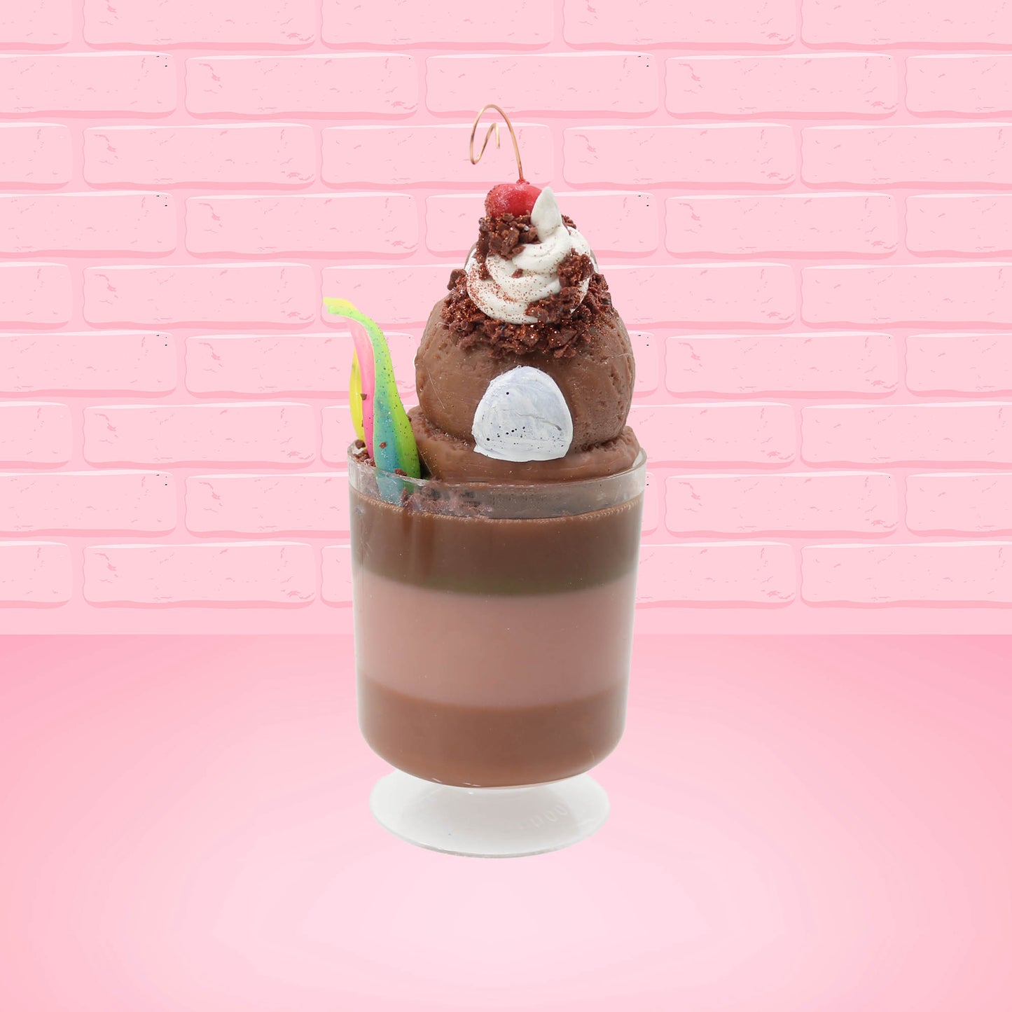 Cute kawaii bunny tall brown and light brown ice cream sundae with brown chocolate crumbles over the bunny, with a cherry on top of the whipped cream swirl.  Three bendy gummy worms are in the dish on the side. This view is from the back.